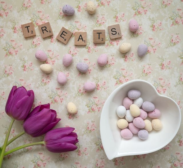 Top view of a delicate Easter arrangement featuring pastel-colored candy eggs, a white heart-shaped bowl, and purple tulips. Wooden letters spelling 'treats' add an elegant touch. Suitable for Easter greetings, spring-themed promotions, and festive decor inspiration.