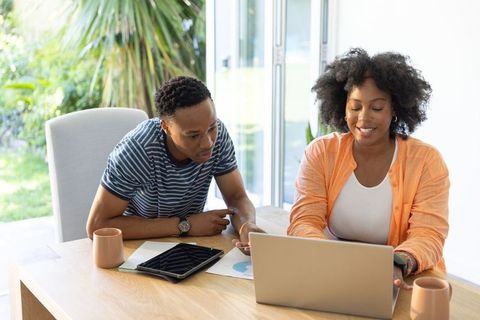 African american couple working from home using laptop and tablet at sunlit table