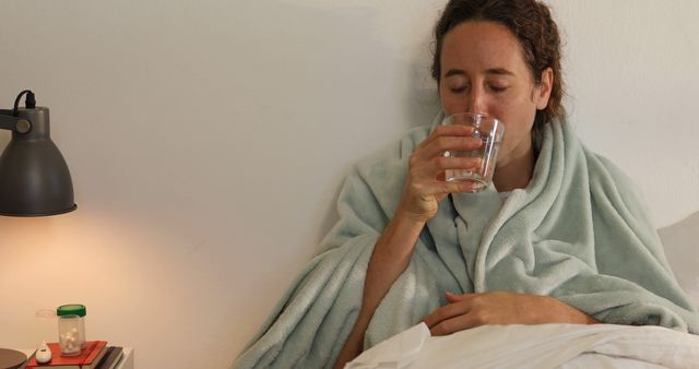 Woman wrapped in a blanket sipping water while resting in bed. Depicts themes of illness, recovery, comfort, and self-care. Useful for articles or content related to health, wellness, home remedies, and medical advice.