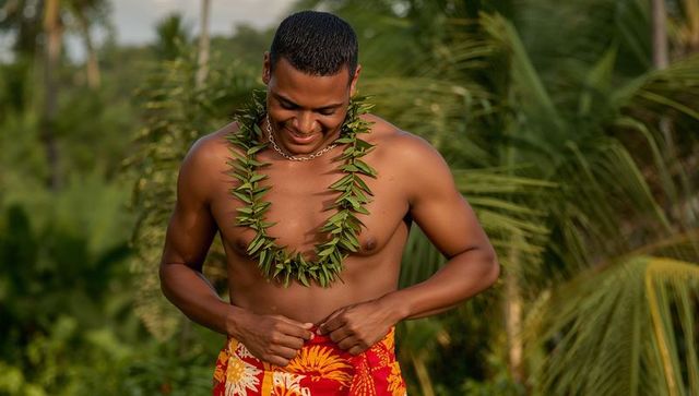 Shirtless asian man adjusting red floral sarong with leaf lei in tropical palm garden