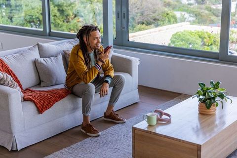 African american man speaking into smartphone on modern sofa by large window with daylight