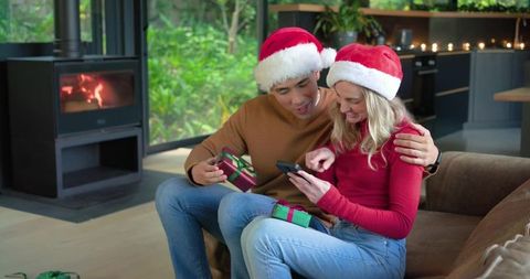 Diverse couple wearing santa hats exchanging gifts and checking phone by cozy fireplace
