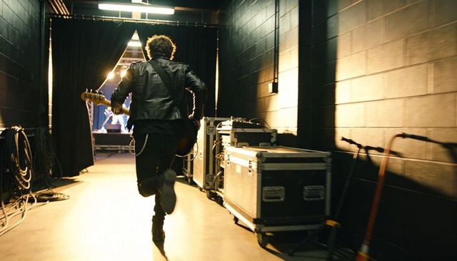 Bassist running backstage toward stage in leather jacket carrying bass guitar under spotlight