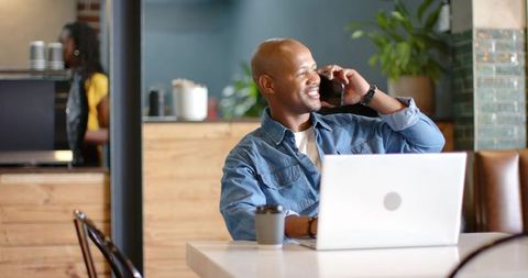 Smiling man talking on smartphone while working on laptop in modern coffeehouse