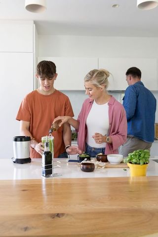 Family preparing green smoothie at bright modern kitchen island, parents guiding teen