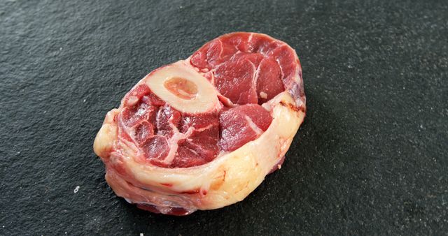 A raw beef shank with a visible marrow bone is placed on a dark textured surface, with copy space. Its rich red color indicates freshness, ideal for culinary preparations like braising or slow cooking.