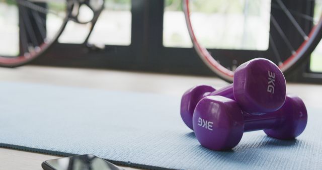 Purple dumbbells resting on a blue yoga mat in a home gym. In the background, part of a bicycle is visible, suggesting a well-equipped fitness space. Ideal for articles on home workouts, fitness routines, or promoting gym equipment.