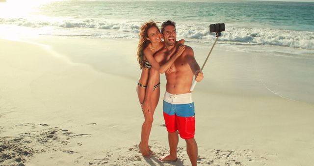 Couple wearing swimwear taking a selfie on sandy beach beside ocean. Woman in bikini and man in swim trunks enjoying summer vacation. Perfect for travel agency marketing, holiday brochures, romantic getaway promotions, and social media content emphasizing vacations and leisure time.