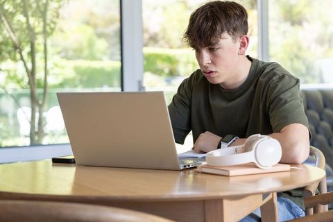 Teenager studying on laptop at home with headphones remote learning in sunlit kitchen