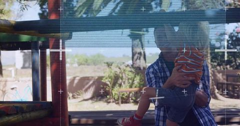 Father holding young son beneath sunlit playground mesh, backyard bonding moment