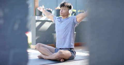 African american man meditating with headphones on balcony yoga mat in sunlight