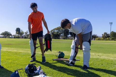 Two teammates fastening cricket pads and preparing protective gear on field during practice