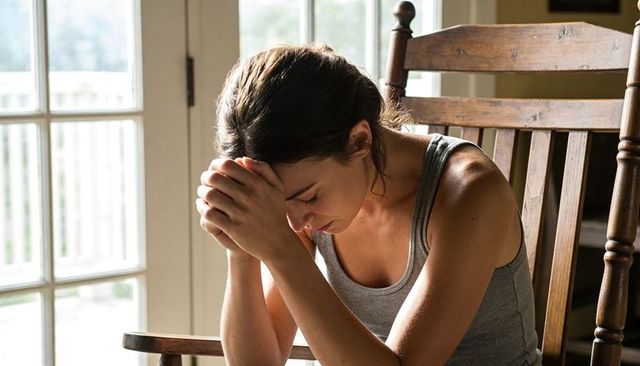 Young woman leaning in rocking chair clasping forehead near glass doors sunlit room contemplation