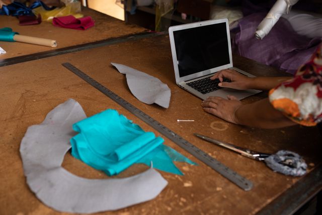 Woman using laptop in a hat factory, surrounded by fabric scraps, scissors, and measuring tape. Ideal for content related to fashion design, handmade crafts, small business, creative workspaces, and artisan industries.