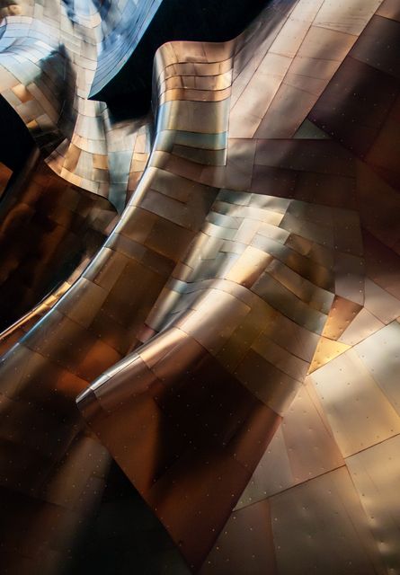 The photograph showcases a close-up of an abstract metallic surface with warm copper and bronze tones, enhanced by dynamic lighting and shadows. The flowing, wavy pattern created by the textured metal panels suggests movement and complexity, creating a visually intriguing and aesthetically pleasing image. This picture is perfect for use in design projects, architectural studies, modern art exhibitions, backgrounds for digital presentations, or as a compelling addition to home or office décor.