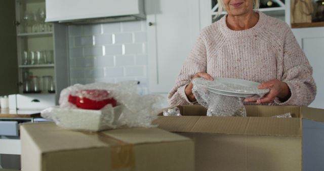 Senior woman packing dishes in cardboard boxes in kitchen. Ideal for use in marketing for moving services, senior living arrangements, or family relocation concepts. Images of packing and wrapping convey preparation, organization, and new beginnings.