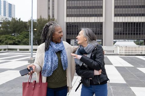 Diverse women friends walking arm-in-arm laughing in city plaza carrying red tote bag