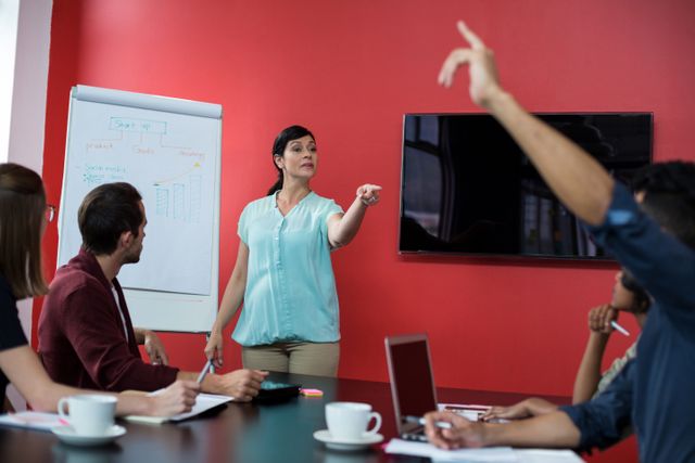 Business professionals engaged in a strategic discussion around a table in a modern office. A woman is leading the meeting, pointing at a flip chart with graphs, while a colleague raises a hand to contribute. Ideal for illustrating corporate teamwork, leadership, business strategy sessions, and collaborative work environments.