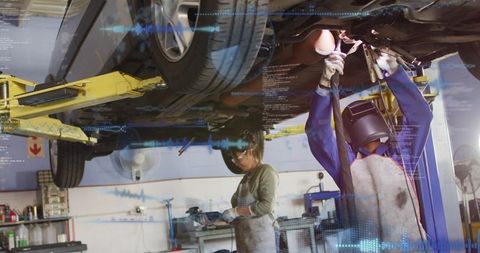 Mechanic wearing welding helmet repairing undercarriage under lift in busy auto workshop