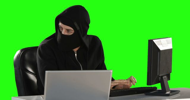 A person in a black hoodie and face mask is engaged in computer work against a green screen background, with copy space. Their attire and the setup suggest a theme related to cybersecurity or hacking.