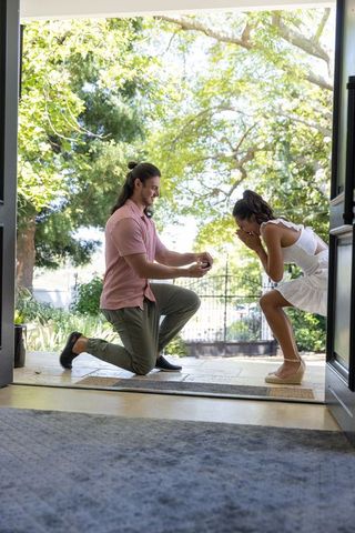 Romantic doorstep proposal man kneeling with ring box woman in white dress reacting