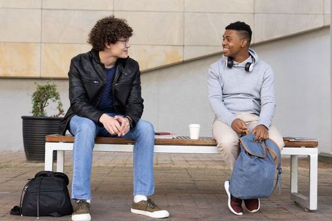 College friends chatting on campus bench sharing coffee and backpacks
