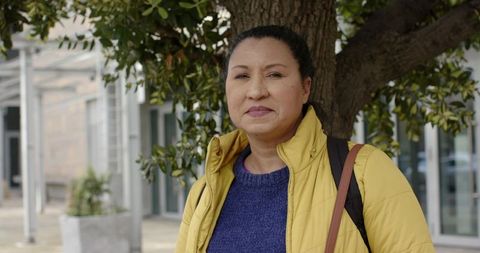 Mature woman standing by tree wearing yellow jacket and backpack in urban plaza
