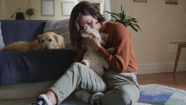 Woman sitting on floor next to sofa cuddling fluffy dog, with another dog lounging nearby. This warm and intimate moment captures a sense of tranquility and companionship. Perfect use for content focusing on pet ownership, emotional well-being, self-care, and home leisure activities.