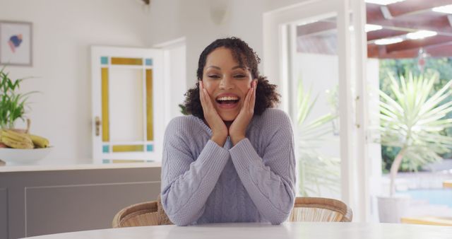 Woman with curly hair is smiling and holding her face while sitting at kitchen table. Table is clean, kitchen has modern design with natural light filling the room. Perfect for depicting themes of happiness, domestic life, and casual moments at home. Ideal for advertisements featuring family life, online blogs about home decor or lifestyle, and social media content about daily living.