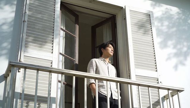 Asian man standing on sunlit balcony looking outward through french doors with shutters