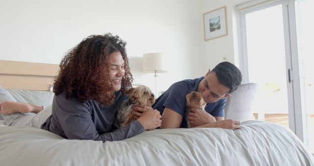 Happy couple playing with their dogs on a comfortable bed in a bright bedroom. They appear relaxed and are smiling, creating a warm and cheerful atmosphere. Perfect for themes related to domestic life, pet ownership, and relaxed home environments.