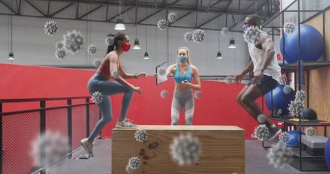 Masked fitness trainer guiding athletes doing plyometric box jumps in industrial gym