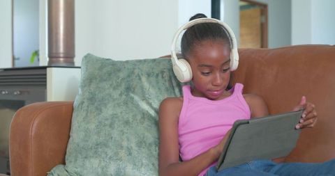 African american preteen girl using tablet wearing headphones on sofa for remote learning