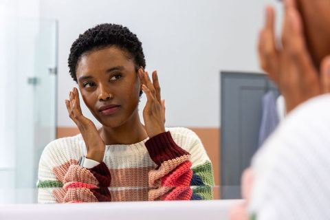 African american woman examining face in mirror wearing striped knit sweater and smartwatch