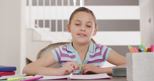 Young girl in casual wear sitting at desk writing and doing homework in bright, modern home environment. Perfect for educational materials, homeschooling content, study guides, family life, or child-focused publications.