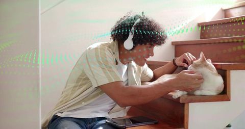 Smiling young man petting cream cat on wooden stairs wearing headphones, cozy home scene