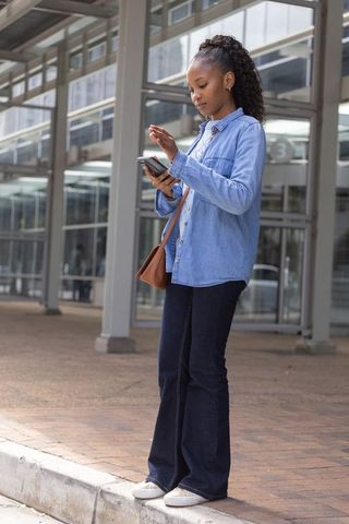 African woman standing and scrolling smartphone by urban glass entrance with crossbody bag
