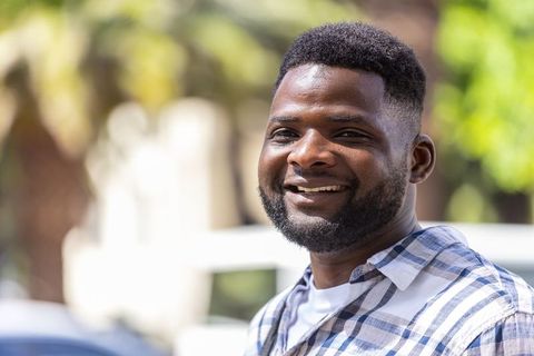 Smiling bearded man standing on sunlit urban street wearing blue plaid shirt and white tee