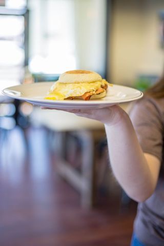 Hand Holding Plate Serving Melted Egg and Ham English Muffin Breakfast Sandwich in Cafe