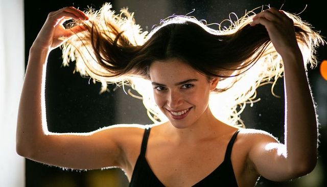 Backlit young woman lifting long hair, smiling rim-lit studio portrait with bokeh dust glamour