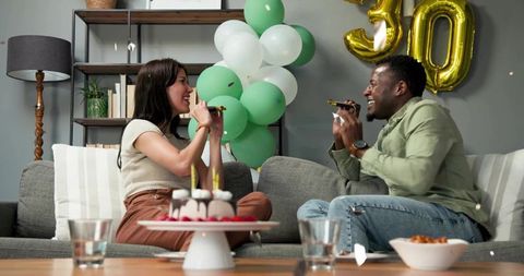 Laughing couple celebrating 30th birthday at home on sofa with cake and balloons