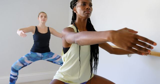 Women engaging in ballet barre workout in fitness studio. One woman with braids wears a sleeveless top and performs squats while another woman in colorful leggings and a tank top follows along. Ideal for content about fitness routines, dance workouts, women's fitness, and strength training exercises.