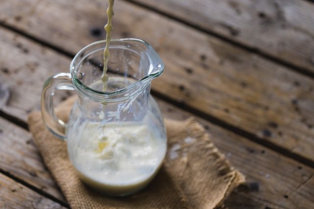 High angle view of fresh milk being poured into a glass jug on a rustic wooden table with a burlap cloth. Ideal for use in food and drink blogs, healthy eating promotions, dairy product advertisements, and farmhouse-themed decor.