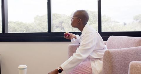 African american clinician taking break in lounge holding apple, glancing out window