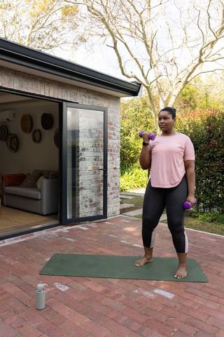 African american woman exercising with dumbbells on backyard brick patio during home workout