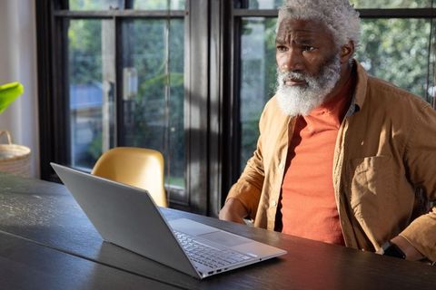 Thoughtful senior african american man working from home on laptop by sunlit window