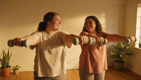 Two women lifting light dumbbells during sunlit home workout, smiling and encouraging