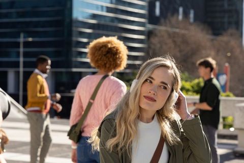 Blonde woman adjusting over-ear headphones in sunlit city plaza with diverse passersby
