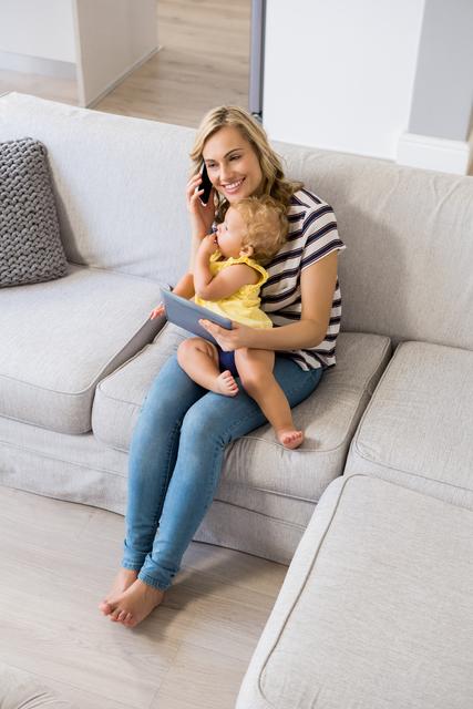 Mother sitting on couch, talking on mobile phone while holding baby girl. Ideal for parenting blogs, family lifestyle articles, and advertisements related to communication technology or home life.