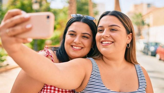 Smiling friends taking selfie on sunny urban street wearing striped tank and gingham dress
